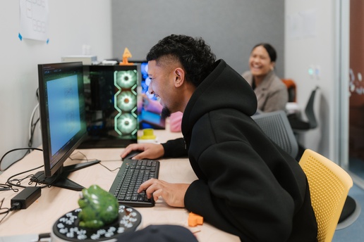 A person in a black hoodie leans toward a computer monitor while typing at a desk with a keyboard and mouse. A lit computer tower with glowing fans sits beside the monitor, and another person works at a desk in the background.