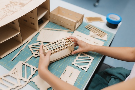 A person assembles a small wooden model on a table covered with cutting mats. Laser‑cut wooden pieces, tape, and a partially built structure are spread out as the person fits patterned panels into place. Provide your feedback on BizChat