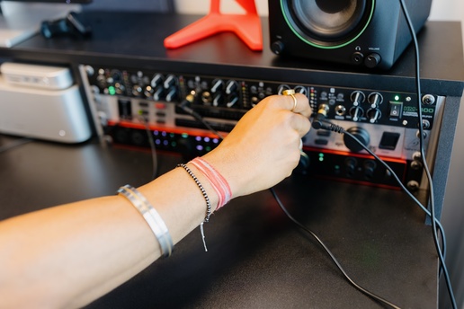 A close-up of a hand adjusting knobs and plugging cables into an audio interface on a desk, with a studio monitor and other recording equipment nearby.