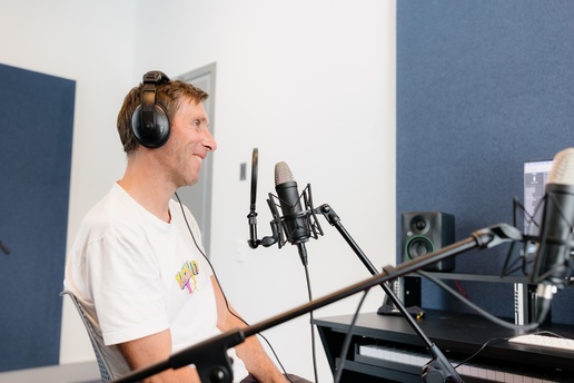 A person wearing headphones sits at a desk in a soundproofed studio, speaking into a mounted microphone with audio equipment, a monitor, and speakers arranged around them.