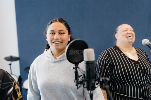 Two people stand in front of microphones in a recording or rehearsal space with a dark blue wall. Music equipment, including a pop filter and a drum set in the background, indicates they are preparing to record or perform.