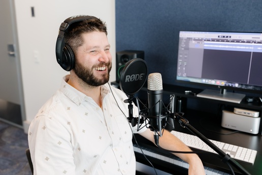 A person wearing headphones speaks into a RØDE microphone at a desk with a monitor showing audio waveforms, seated in a studio-style space with a blue acoustic panel behind them
