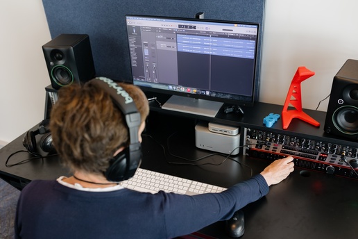 A person wearing headphones works at a desk with a digital audio workstation on a large monitor, surrounded by studio speakers, an audio interface, a keyboard, and a small red guitar-shaped object