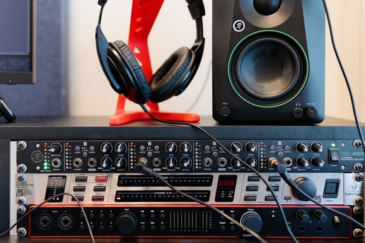 A pair of black over‑ear headphones rests on a red stand beside a studio monitor speaker and rack‑mounted audio gear with knobs, buttons, and connected cables.