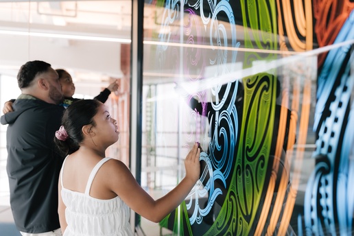 Two people interact with a large, colorful digital display filled with swirling patterns in green, blue, and orange. One person reaches toward the screen while another stands nearby, both engaging with the artwork.