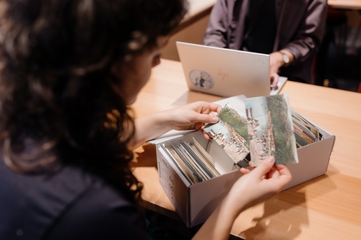 A person sits at a wooden table examining old photographs from a box filled with prints and documents. Another person sits across the table using a laptop. The setting appears to be an archive, library, or workspace.