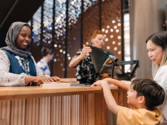 A staff member stands behind a wooden counter assisting a visitor and a child who is reaching toward the counter. Additional people are visible in the background near decorative panels with circular cut‑out patterns.