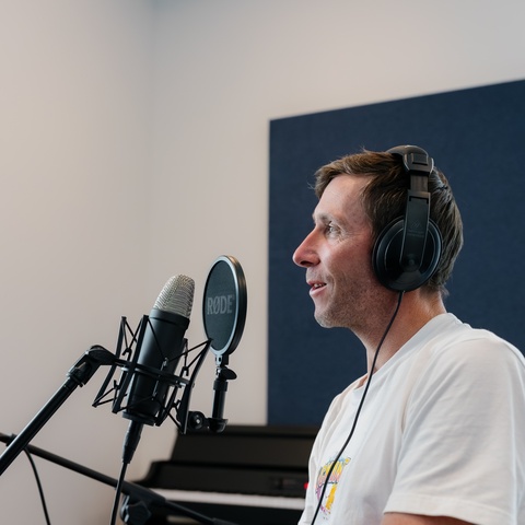 A person wearing headphones sings or speaks into a large condenser microphone with a RØDE pop filter, standing near a piano in a studio with a blue acoustic panel behind them.