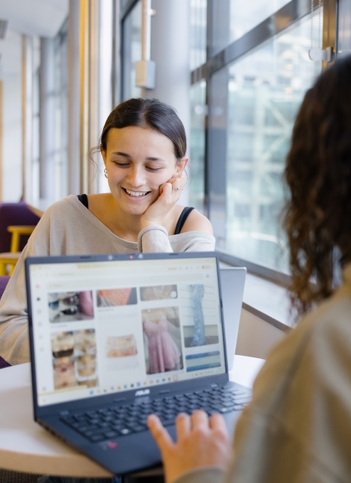 A person sits at a table in a bright space with large windows, looking toward another person using a laptop. The laptop screen shows a grid of images, and bookshelves and seating are visible in the background.