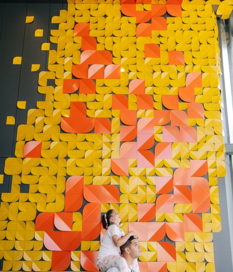 A person carries a child on their shoulders while standing in front of a tall wall covered in a large geometric art installation made of yellow, orange, and pink folded shapes arranged in a mosaic-like pattern.
