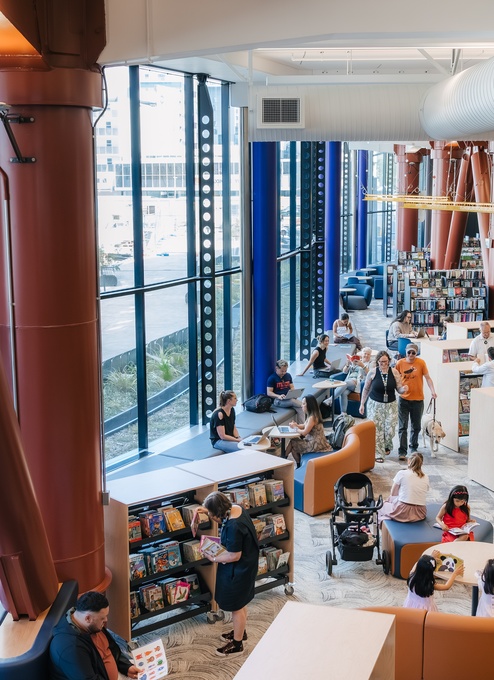 A spacious, modern library filled with bookshelves, seating areas, and people reading, browsing, and talking. Large floor‑to‑ceiling windows line one side, with tall structural columns adding color and height to the open interior