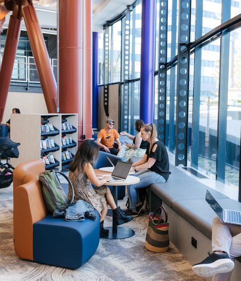 People work and relax in a bright library lounge with large windows, blue and orange seating, and tall structural columns. Some sit with laptops or books, while others talk or move through the space near bookshelves and strollers.