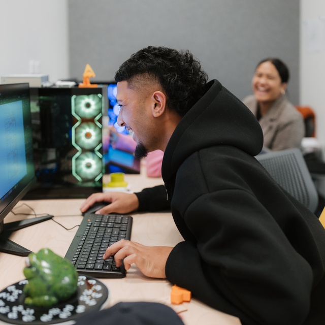 A person in a black hoodie leans toward a computer monitor while typing at a desk with a keyboard and mouse. A lit computer tower with glowing fans sits beside the monitor, and another person works at a desk in the background.