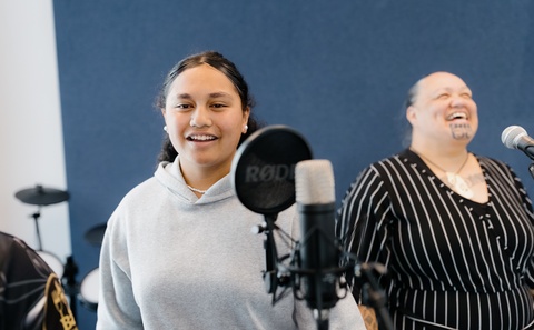 Two people stand in front of microphones in a recording or rehearsal space with a dark blue wall. Music equipment, including a pop filter and a drum set in the background, indicates they are preparing to record or perform.
