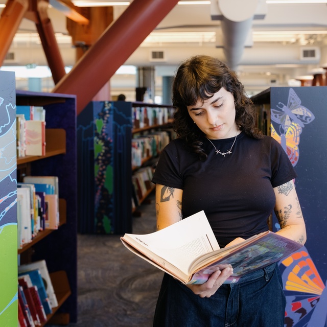 A person stands in a library aisle reading an open book. Colorful panels with illustrated designs frame the shelves, and rows of books extend into the background beneath exposed beams and bright overhead lighting.
