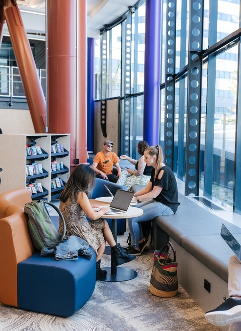 People work and relax in a bright library lounge with large windows, blue and orange seating, and tall structural columns. Some sit with laptops or books, while others talk or move through the space near bookshelves and strollers.