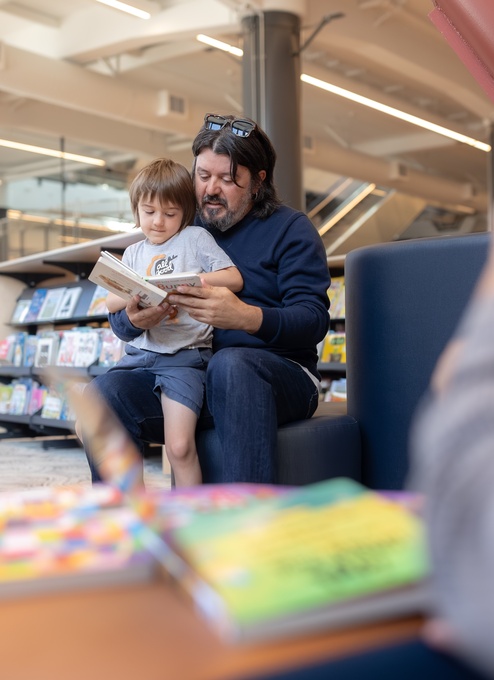 An adult sits on a library chair reading a book with a child on their lap. Bookshelves and displays fill the background, while another person is visible in the foreground, slightly out of focus.