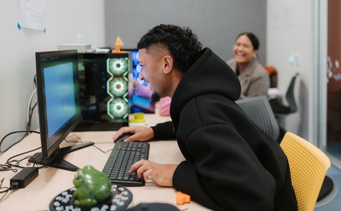 A person in a black hoodie leans toward a computer monitor while typing at a desk with a keyboard and mouse. A lit computer tower with glowing fans sits beside the monitor, and another person works at a desk in the background.