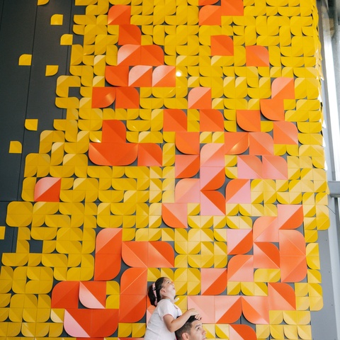 A person carries a child on their shoulders while standing in front of a tall wall covered in a large geometric art installation made of yellow, orange, and pink folded shapes arranged in a mosaic-like pattern.