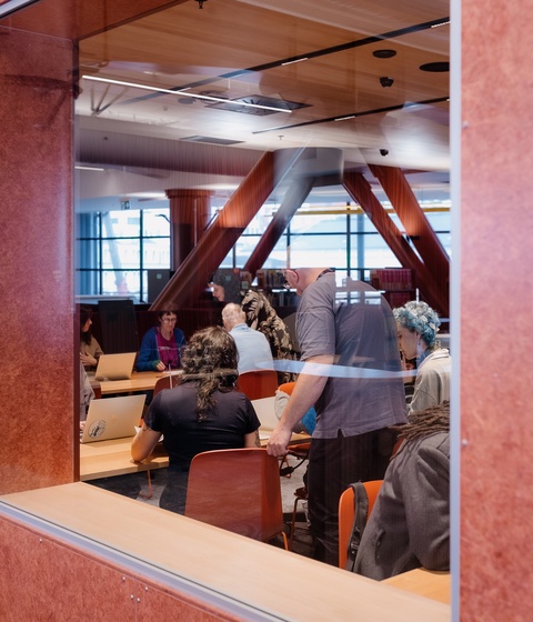 A view through a large interior window into a classroom or meeting area where people sit at tables working on laptops and papers. The bright room features tall structural beams and wide windows in the background.
