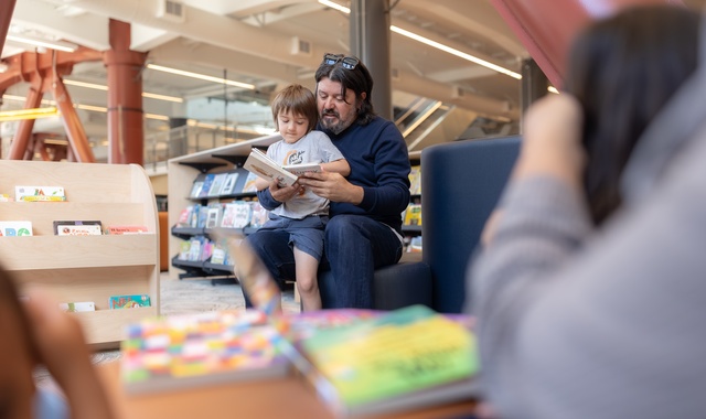 An adult sits on a library chair reading a book with a child on their lap. Bookshelves and displays fill the background, while another person is visible in the foreground, slightly out of focus.