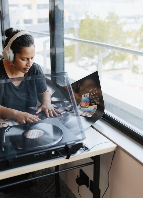 A person wearing headphones sits at a table beside large windows, listening to a vinyl record on a turntable. An open record sleeve rests next to the player in a bright, modern seating area.