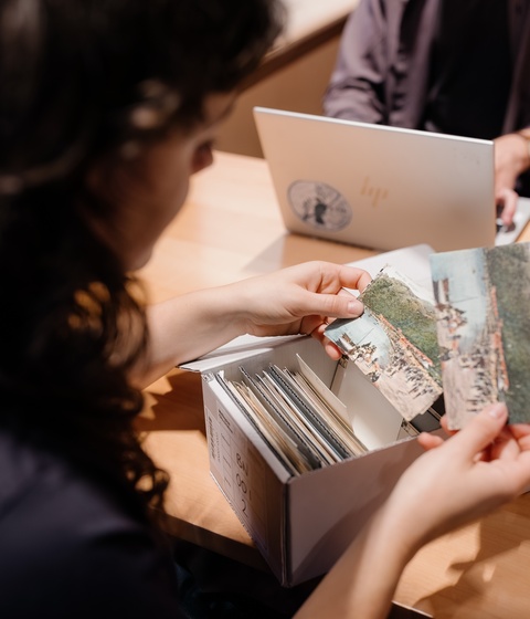 A person sits at a wooden table examining old photographs from a box filled with prints and documents. Another person sits across the table using a laptop. The setting appears to be an archive, library, or workspace.