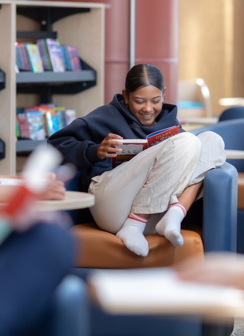 A person sits curled up on a cushioned seat in a library, reading a book while holding it close. Bookshelves filled with colorful titles line the background, and other people are seated and reading nearby, slightly out of focus.
