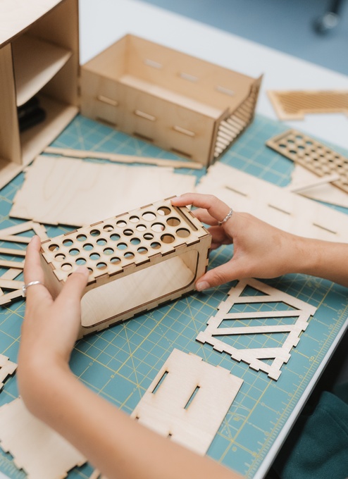 A person assembles a small wooden model on a table covered with cutting mats. Laser‑cut wooden pieces, tape, and a partially built structure are spread out as the person fits patterned panels into place. Provide your feedback on BizChat