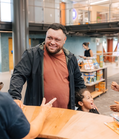 People gather around a wooden counter inside a library or community space. Bookshelves, structural columns, and an upper walkway are visible in the background, while children’s books and a person’s outstretched hand are in the foreground.