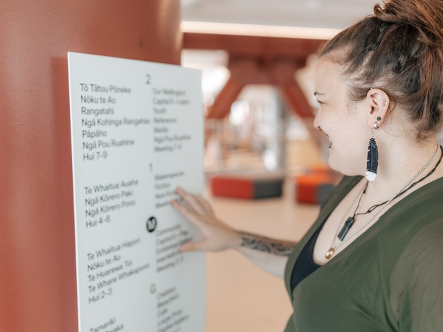 A person stands beside a large red column, pointing at a bilingual English–Māori sign in an indoor public space with beams and seating visible in the background.