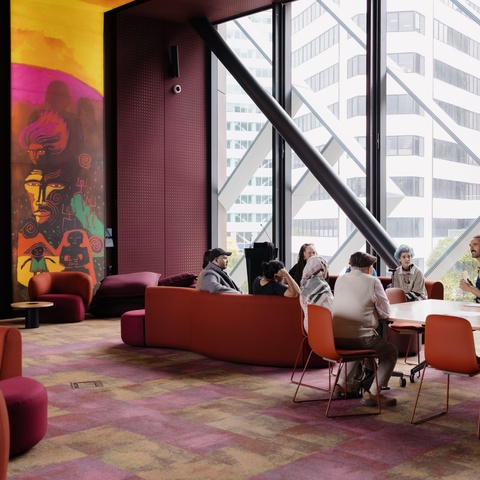 group of people sit around tables and lounge seating in a brightly lit room with large floor‑to‑ceiling windows. Bold, colorful wall art featuring abstract figures fills one side of the space, which is furnished with red and orange chairs and couches.