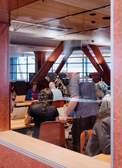 A view through a large interior window into a classroom or meeting area where people sit at tables working on laptops and papers. The bright room features tall structural beams and wide windows in the background.
