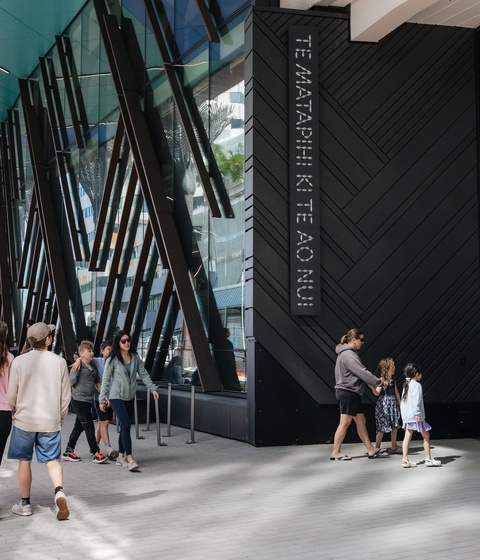 People walk past the entrance of a modern building with tall angled beams and large glass panels. A sign reading “TE MATAPIHI KI TE AO NUI” is mounted beside the doorway as groups and families move along the wide outdoor walkway.