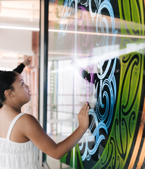 Two people interact with a large, colorful digital display filled with swirling patterns in green, blue, and orange. One person reaches toward the screen while another stands nearby, both engaging with the artwork.