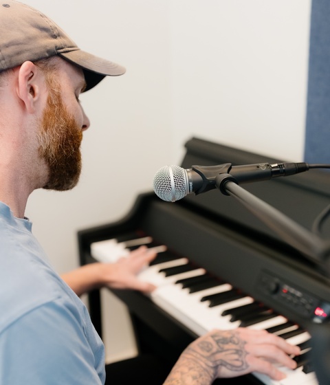 A person in a light cap and blue shirt plays a black upright piano with a microphone positioned nearby in a simple indoor studio with white walls and a blue acoustic panel.