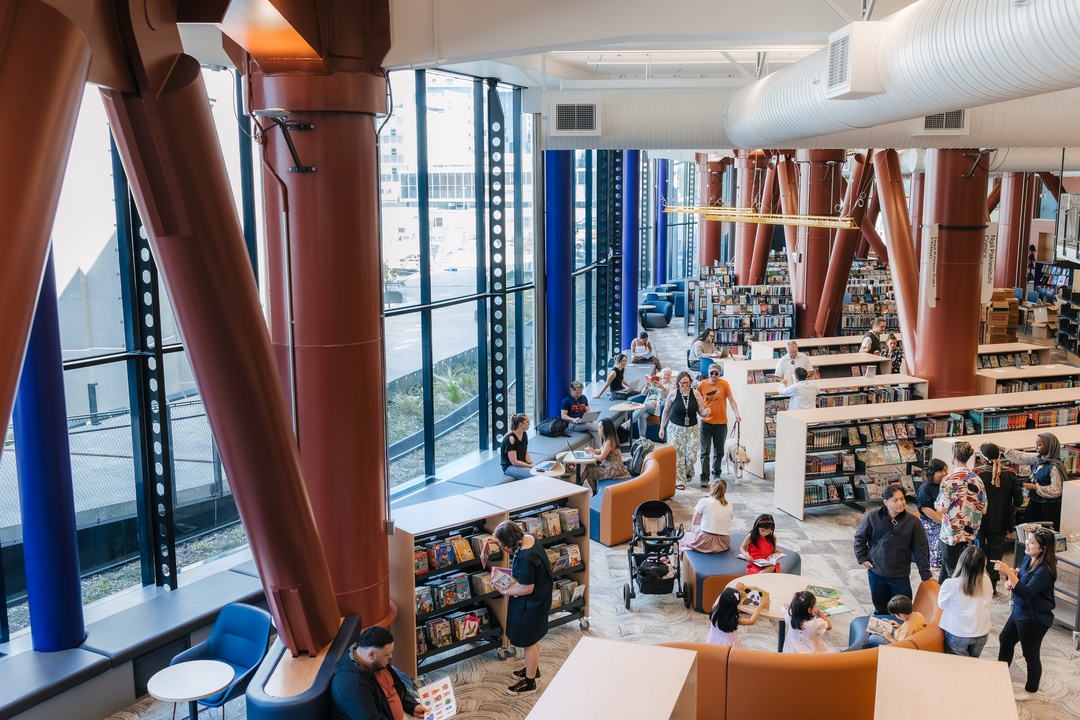 A spacious, modern library filled with bookshelves, seating areas, and people reading, browsing, and talking. Large floor‑to‑ceiling windows line one side, with tall structural columns adding color and height to the open interior
