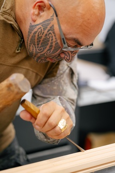A person uses a chisel and wooden mallet to carve a piece of wood in a workshop, focused intently on their craft.