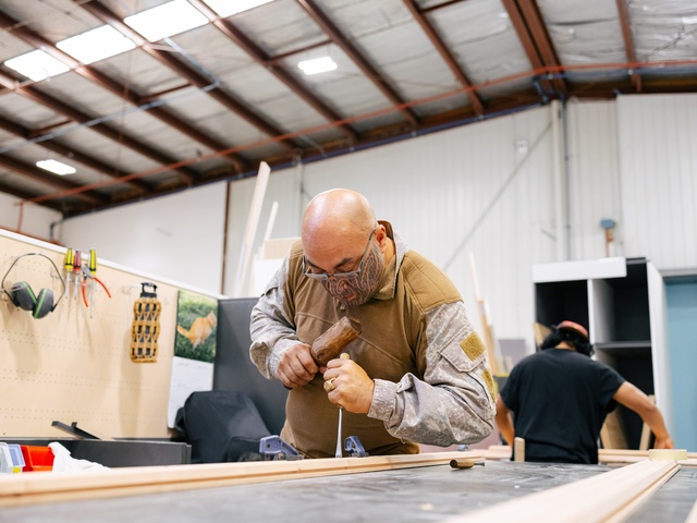 A person works on a piece of wood in a workshop, using a mallet and chisel at a workbench. Tools hang on a pegboard in the background, and another person works at a separate station in the spacious, industrial workspace.