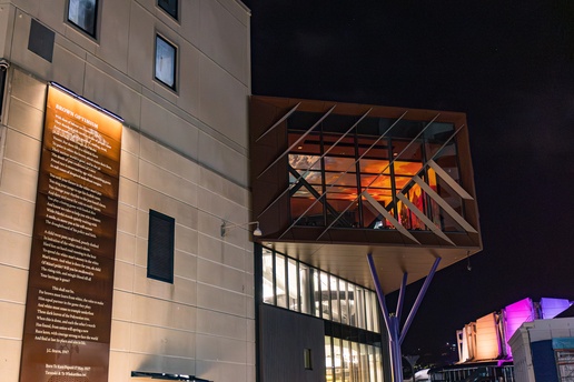 A modern building at night features a cantilevered glass section lit from within, supported by angled columns, with a tall illuminated vertical panel on the exterior wall.