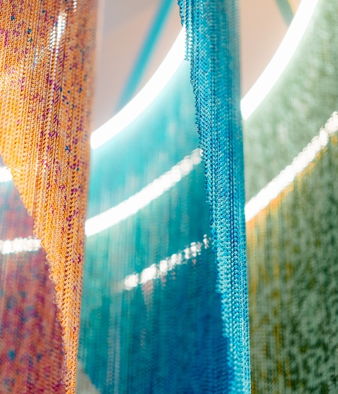 Colorful hanging strands of beaded or textured material in yellow, orange, blue, and green cascade vertically, illuminated by circular lights above to create a soft, glowing display.