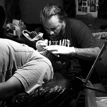 A tattoo artist wearing gloves works on the back of a person lying on a padded surface. The scene is in a dimly lit studio with equipment, artwork, and supplies visible in the background.